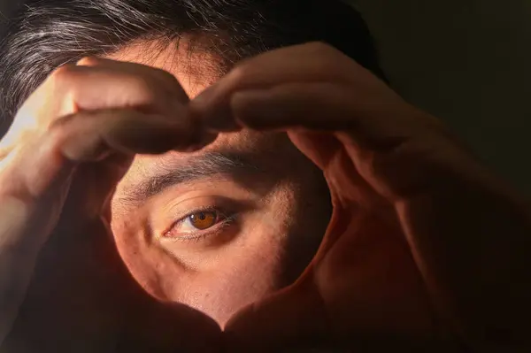 Bir adam gözlerini çevreleyen bir kalp şekli oluşturmak için ellerini kullanıyor. Oyuncu ama anlamlı bir jest sergiliyor. Sıcak ışık onun yüz hatlarını ön plana çıkarıyor ve jestin arkasındaki duyguya dikkat çekiyor..