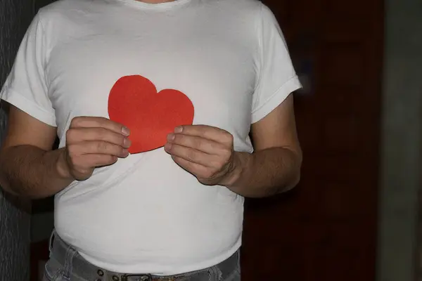 Kırmızı bir kalp sembolü taşıyan beyaz tişörtlü bir adamın yakın plan çekimi, duygu, aşk ve bağlantı temalarını iletiyor..