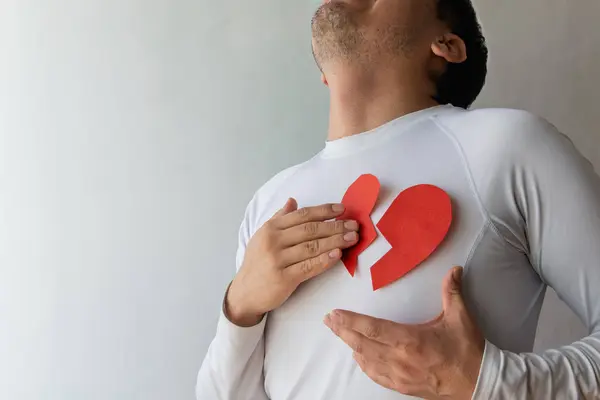 Tarafsız bir arkaplanda duran bir adam kırmızı kağıt bir kalbi göğsünün ortasına bir yırtık ile yerleştiriyor ve derin duygusal acıyı işaret ediyor. Yüzündeki ifade üzüntü ve yansımayı ortaya çıkarıyor, kalp kırıklığıyla başa çıkma mücadelesini yakalıyor.