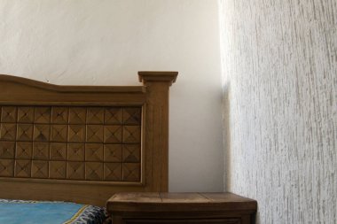 Karmaşık oymalı ahşap bir yatak çerçevesi bu rahat yatak odasında dikkatleri çekiyor. Açık renkli duvar ince bir dokuyu gösterirken, basit bir ahşap komodin yatağın yanında duruyor. Doğal ışık boşluğu nazikçe doldurur, sıcak ve davetkar bir ortam yaratır.