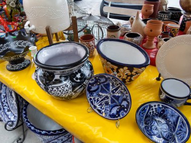 Parlak renkli Talavera çömlekleri Meksika 'daki hareketli bir pazarda sarı bir masada sergileniyor. Zanaatkarlar, bu geleneksel sanat eserleriyle ilişkili zengin kültür mirasını ve zanaatkarlığını vurgulayarak karmaşık bir şekilde tasarlanmış kâseler, tabaklar ve kavanozlar sergiliyorlar.