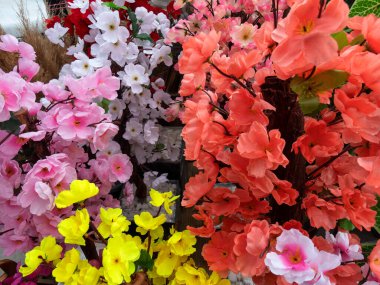 Canlı bir suni çiçek sergisi market tezgahını bir dizi renkle dolduruyor. Pembe, turuncu, sarı ve beyaz çiçekler çarpıcı bir görüntü oluşturur. Çiçekler dikkatlice düzenlenmiş, canlı görünüşlerini sergiliyorlar..