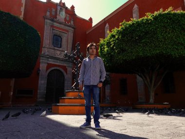 Santiago de Queretaro 'nun tarihi merkezinde kendine güvenen genç bir adam duruyor. Etrafında güzel manikürlü ağaçlar ve çarpıcı bir halk heykeli var. Bir güvercin sürüsü de kaldırım taşlarını araştırıyor..