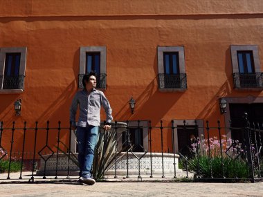 Santiago de Queretaro Tarih Merkezi 'nde genç bir birey süslü demir bir çitin karşısında gelişigüzel duruyor. Arkaplanda, bölgenin tipik mimari unsurlarının sergilendiği çok sayıda pencereli çarpıcı turuncu bir bina yer alıyor..