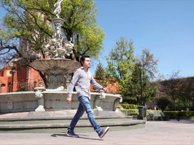 Santiago de Queretaro Tarih Merkezi 'ndeki güzel bir çeşmenin yanından güvenle geçen genç bir adam görülüyor. Yeşillik bölgeyi çevreleyerek, sakin yürüyüşler için canlı bir zemin oluşturuyor..