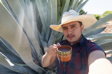 Meksikalı bir adam, Meksika 'nın güneşli bir bölgesinde yüksek bir agave bitkisinin yanında dururken geleneksel bir içecek olan pulkanın tadını çıkarıyor. Geniş kenarlı bir şapka ve ekose gömlek giyiyor, elinde içecekle dolu dekoratif bir bardak tutuyor. Zengin sörfçü.