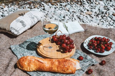 Summer romantic rustic picnic on the coast with white wine glass, baguette, cherries, grape, open book, pillow. Sea beach. Vacation, rest in Europe. Alcohol drink. Inspiration. High quality photo