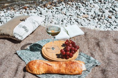 Summer romantic rustic picnic on the coast with white wine glass, baguette, cherries, grape, open book, pillow. Sea beach. Vacation, rest in Europe. Alcohol drink. Inspiration. High quality photo