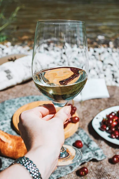 Hand with white wine glass. Summer romantic rustic picnic on the coast, bottle, baguette, cherries and grape. Sea beach. Vacation, rest in Europe.