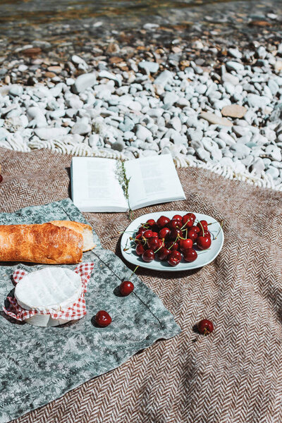 Summer romantic rustic picnic on the plaid. French style. Baguette, cherries, camembert cheese, open book from above. Vacation, rest on the beach alone.