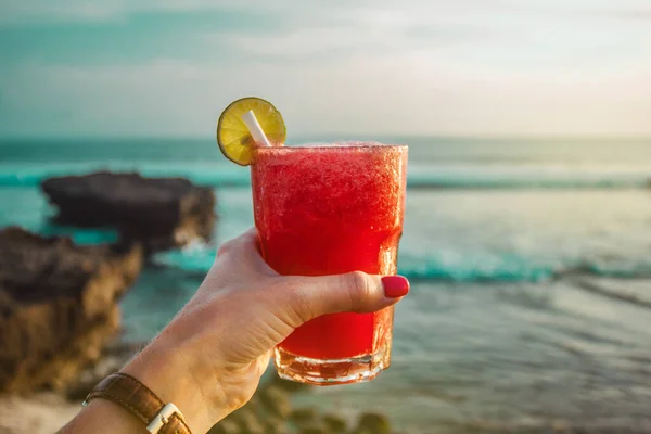 Hand with glass of cocktail with watermelon juice on the background of the ocean. Sunset on the sea. Healthy red drink. Bali, Indonesia, Canggu, Echo beach. Vacation time