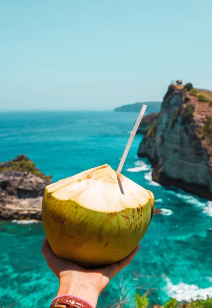 A hand with fresh coconut on the ocean and cliffs tropical background. Tropical landscape. Vacation mood. Bali, Indonesia