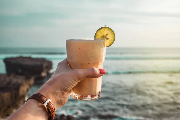 Hand with glass of cocktail on the background of the ocean. Sunset on the sea. Healthy white drink. Bali, Indonesia, Canggu, Echo beach. Vacation time