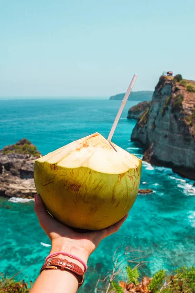 stock image A hand with fresh coconut on the ocean and cliffs tropical background. Tropical landscape. Vacation mood. Bali, Indonesia