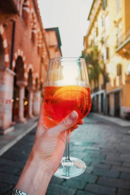 Eski binaların arka planında bir bardak turuncu kokteyl spreyli kadın eli. Alkollü içecek ve buz. İtalya, Verona 'da güneşli bir yaz günü. Avrupa 'da Tatil.