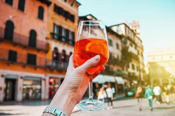 Eski binaların arka planında bir bardak turuncu kokteyl spreyli kadın eli. Alkollü içecek ve buz. İtalya, Verona 'da güneşli bir yaz günü. Avrupa 'da Tatil.
