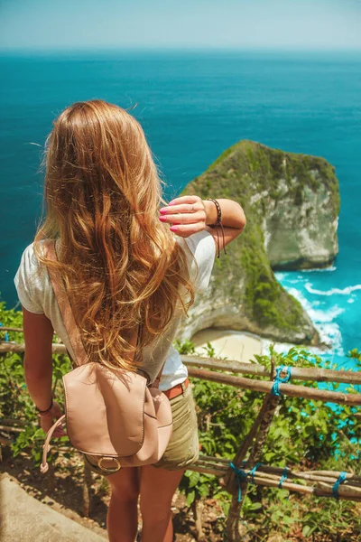 stock image Travel girl with backpack in shorts on the ocean, cliffs and tropical Kelingking beach background. Nusa Penida, Indonesia. Female silhouette, blonde hair.