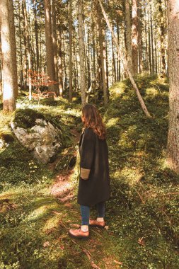 Uzun paltolu kahverengi saçlı genç bir kadın sık bir ormanın ortasında tek başına yürüyor. Atmosferik dikey fotoğraf yeşil tonlarda