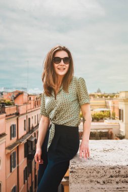 Güneş gözlüklü genç bir kadın panoramik Roma manzaralı bir çatıda oturuyor. İtalya 'da tatilde olan bir kız. Güney Avrupa 'da güneşli bir gün. Ünlü bir yerin panoramik manzarası. Sosyal için dikey fotoğraf