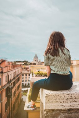 Genç bir kadın sırtı çatıda, Roma 'nın panoramik manzarasıyla oturuyor. İtalya 'da tatilde olan bir kız. Güney Avrupa 'da güneşli bir gün. Ünlü yerin panoramik görünümü.