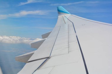 Bir uçak penceresinden mavi gökyüzü ve beyaz bulutlu bir uçak kanadı görünüyor.