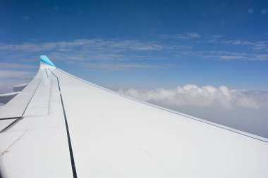 Bir uçak penceresinden mavi gökyüzü ve beyaz bulutlu bir uçak kanadı görünüyor.