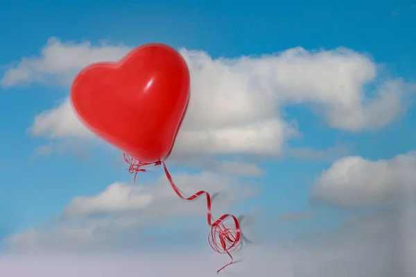 A red heart-shaped balloon with red string is lying on a large picture with a blue sky and white clouds