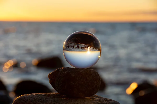 stock image Glass ball lies on the top stone of stacked stones, at sunset on the beach, the sea and the setting sun are reflected in the ball