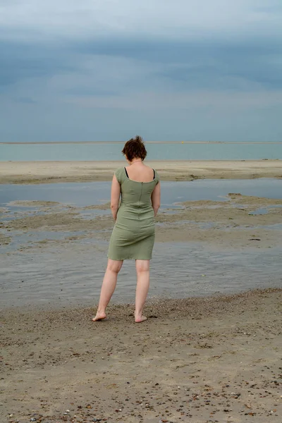 stock image Young woman in a short green dress is standing in front of the sea on a sandy beach