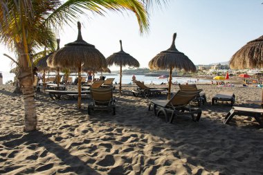 Kosta Adeje, Tenerife, İspanya 'da palmiye ağaçları, deniz, güverte sandalyeleri ve şemsiyeleriyle Torviscas Playa kumlu plajı