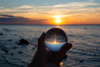 Yaşlı bir insanın eli cam bir top tutuyor, deniz ve batan güneş topa yansıyor