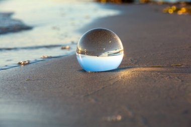 Camdan bir top kumlu sahilde dalgalarda yatıyor, deniz ve batan güneş topa yansıyor.