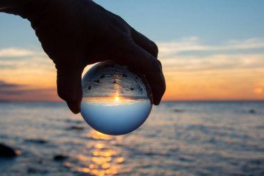 Yaşlı bir insanın eli cam bir top tutuyor, deniz ve batan güneş topa yansıyor