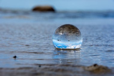 Camdan bir top kumlu sahilde dalgalarda yatıyor, deniz ve batan güneş topa yansıyor.