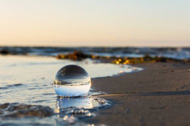 Camdan bir top kumlu sahilde dalgalarda yatıyor, deniz ve batan güneş topa yansıyor.