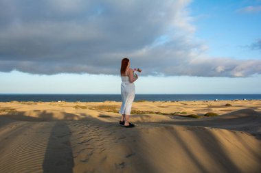 Kum tepelerinde genç bir kadın elinde bir su şişesi, kollarını denize doğru uzatmış duruyor.