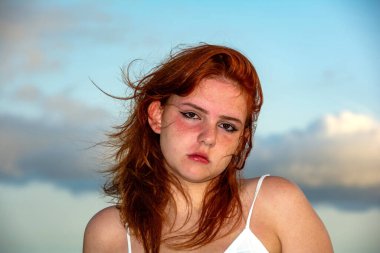 Kızıl saçlı genç bir kızın portresi, mavi gökyüzü ve bulutlar, rüzgarla dalgalanan saçlar