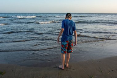 Şort ve kep giyen bir çocuk denizin kıyısındaki kumlu bir sahilde duruyor, mavi bir gökyüzü var.
