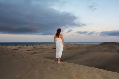 Genç bir kız, kum tepelerinde beyaz bir elbise içinde, öğleden sonra gökyüzü ve bulutlarla duruyor.