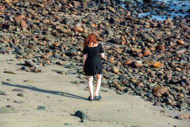 Kısa siyah bir elbise giyen genç bir kız ve taşlı bir plajda arkadan