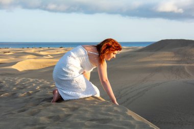 Beyaz elbiseli, genç, kızıl saçlı bir kız öğleden sonra kum tepelerindeki kumlarla oynuyor ve güneşin son ışıklarının tadını çıkarıyor.