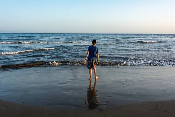 A boy wearing shorts and a cap stands on a sandy beach by the sea, with a blue sky