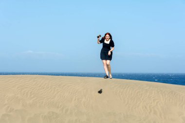 Siyah, kısa yaz elbisesi içinde genç bir kadın kum tepeciğinde duruyor ve mavi gökyüzü ile bir ayakkabısını kaybetmiş.