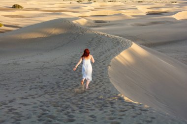 Beyaz elbiseli güzel, kızıl saçlı bir kız öğleden sonra kumulda denize doğru koşuyor.