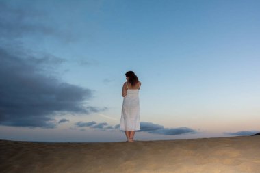 Genç bir kadın sırtı kum tepelerinde duruyor.
