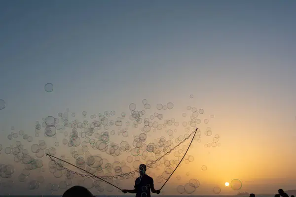 A man in silhouette makes lots of soap bubbles with a soap bubble stick, they float in the orange sky at sunset