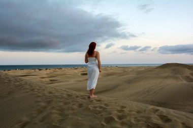 Genç bir kız, kum tepelerinde beyaz bir elbise içinde, öğleden sonra gökyüzü ve bulutlarla duruyor.