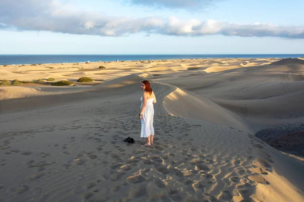 Genç bir kadın kum tepelerinde duruyor, ayakkabıları önünde kumda yatıyor, arka planda deniz var.