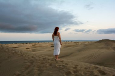 Genç bir kız, kum tepelerinde beyaz bir elbise içinde, öğleden sonra gökyüzü ve bulutlarla duruyor.
