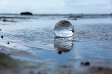 Camdan bir top kumlu sahilde dalgalarda yatıyor, deniz ve batan güneş topa yansıyor.